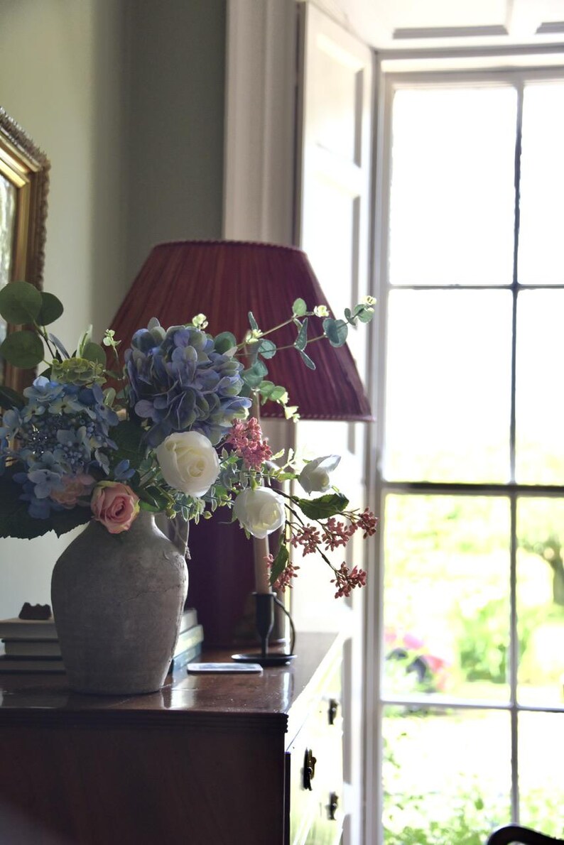Pu&ograve; includere: Una composizione floreale in un vaso grigio con ortensie blu, rose bianche e accenti rosa. Un paralume bordeaux &egrave; dietro il vaso su una superficie in legno scuro. Sullo sfondo una finestra con vista sul verde.