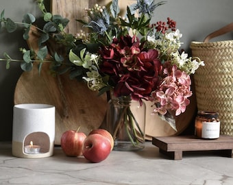 May include: A still life arrangement featuring a vase of burgundy and pink flowers, a lit candle in a ceramic oil warmer, and three red apples. A small candle in a glass jar sits on a wooden tray. A woven basket is in the background.