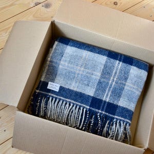 Puede incluir: Una manta de lana a cuadros azules y blancos con flecos est&aacute; doblada y colocada dentro de una caja de cart&oacute;n. La etiqueta en la manta dice "Wool Mill".