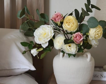 May include: A white ceramic vase with a textured surface and two handles on each side, filled with a bouquet of white, yellow, and pink roses.