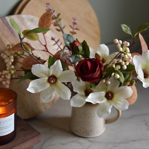 May include: A floral arrangement in a speckled beige mug, featuring white magnolia blooms, burgundy roses, and various greenery. A lit candle in a brown glass jar sits nearby. A wooden cutting board is in the background.