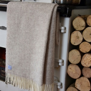 Op de afbeelding: Een beige herringbone deken met witte franjes die aan een wit vintage fornuis hangt met een stapel brandhout rechts.