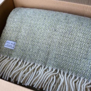 K&ouml;nnte beinhalten: Eine weiche, gr&uuml;ne Wolldecke mit wei&szlig;em Fransenbesatz. Die Decke ist gefaltet und in einem Karton verpackt. Das Etikett auf der Decke lautet "Wool Mill".