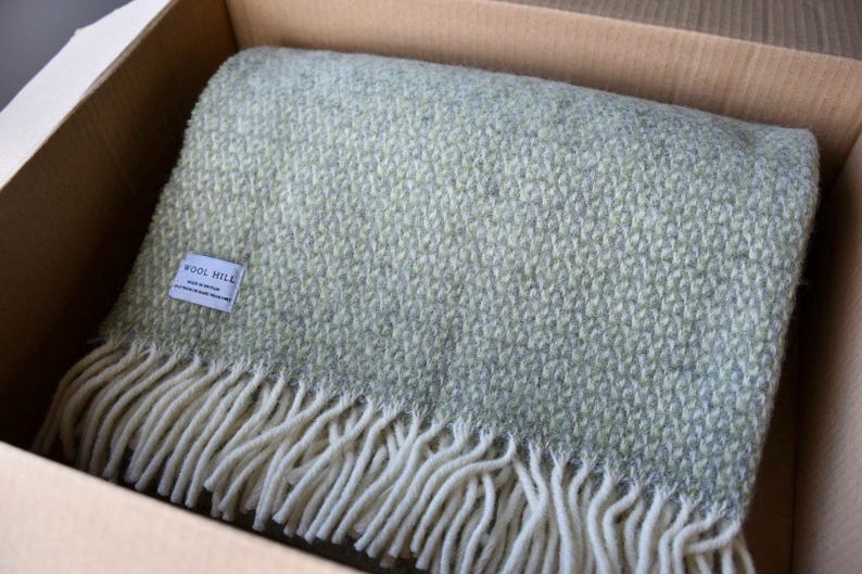K&ouml;nnte beinhalten: Eine weiche, gr&uuml;ne Wolldecke mit wei&szlig;em Fransenbesatz. Die Decke ist gefaltet und in einem Karton verpackt. Das Etikett auf der Decke lautet "Wool Mill".