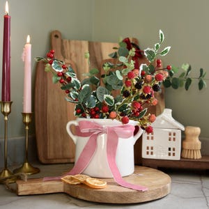 May include: A white ceramic pitcher with a pink ribbon bow, filled with berry and leaf stems. Two lit candles in gold holders and a small white house figurine are also visible. The arrangement sits on a wooden cutting board.