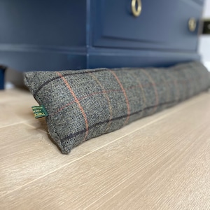 May include: A rectangular door draft stopper made of gray tweed fabric with a red and orange plaid pattern. The door draft stopper is resting on a light-colored wooden floor. A dark blue cabinet is in the background.