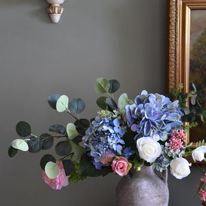 Pu&ograve; includere: Una composizione floreale in un vaso grigio strutturato. Il bouquet presenta ortensie blu, rose bianche, rose rosa e fogliame verde. Il vaso &egrave; su una superficie di legno contro un muro grigio-verde.