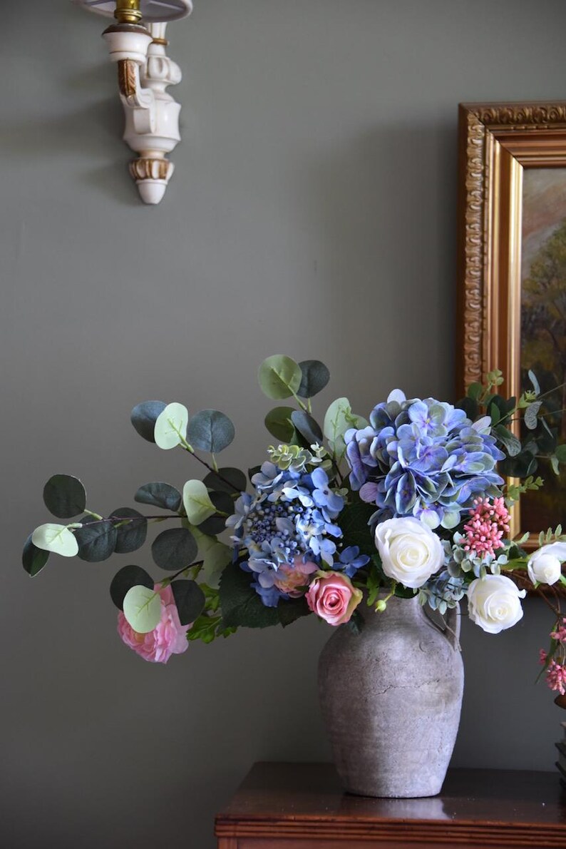 Pu&ograve; includere: Una composizione floreale in un vaso grigio strutturato. Il bouquet presenta ortensie blu, rose bianche, rose rosa e fogliame verde. Il vaso &egrave; su una superficie di legno contro un muro grigio-verde.