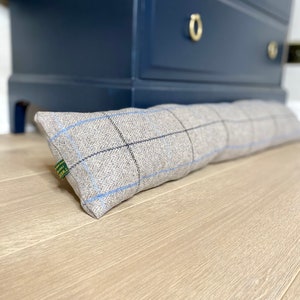 May include: A rectangular, gray tweed door draft stopper with blue and black plaid lines. The door draft stopper is resting on a light wood floor in front of a dark blue dresser with gold hardware. A green tag is attached to the end.