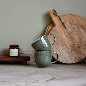 May include: Two green ceramic mugs stacked on a marble countertop. A small jar with a label is on a wooden tray to the left. Wooden chopping boards are in the background. The mugs have a white rim and a handle.