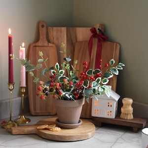 Puede incluir: Una escena de cocina decorativa con una maceta de terracota con un arreglo de bayas y hojas, flanqueada por velas encendidas en candelabros dorados. Tablas de cortar de madera y un pequeño portavelas en forma de casa.