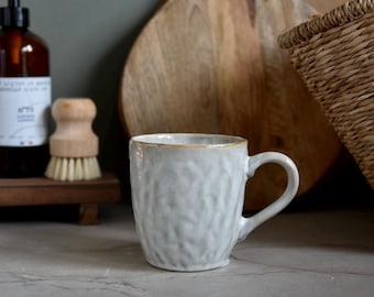 May include: A textured white ceramic mug with a handle, set on a marble surface. The mug has a light brown rim. In the background are wooden cutting boards, a woven basket, a wooden brush, and a brown bottle.