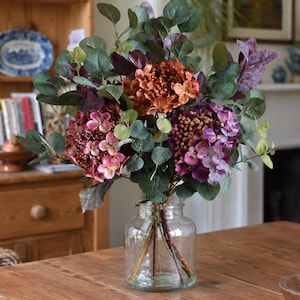Pode incluir: Um vaso de vidro transparente contendo um arranjo floral de flores de hortênsia artificiais em tons de ferrugem, rosa e roxo, acentuado com folhas de eucalipto verdes e roxas escuras. Os caules são visíveis através do recipiente de vidro.