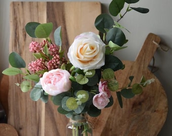 May include: A bouquet of pink and cream roses and eucalyptus leaves in a clear glass vase. The vase is sitting on a wooden cutting board.