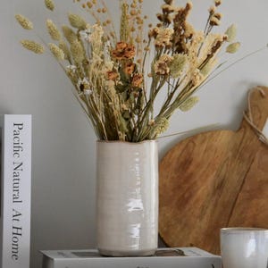 Può includere: Un vaso di ceramica color crema pieno di fiori ed erbe secche. Il vaso è posto su una mensola con libri e una tazza. La composizione comprende elementi beige, marroni e marroni, creando un'estetica naturale e neutra.