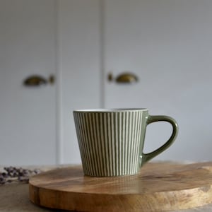 May include: Olive green ceramic mug with vertical white stripes, set on a wooden cutting board. The mug has a curved handle and a white interior. The background features a blurred white cabinet.