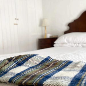 Puede incluir: Una cama blanca con una manta de cuadros azules, verdes y blancos doblada en la parte superior. La cama tiene una cabecera de madera y est&aacute; en un dormitorio con paredes blancas.