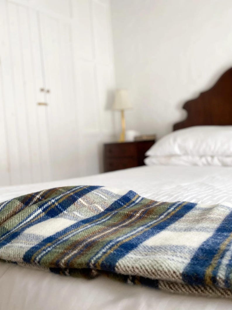 Puede incluir: Una cama blanca con una manta de cuadros azules, verdes y blancos doblada en la parte superior. La cama tiene una cabecera de madera y est&aacute; en un dormitorio con paredes blancas.