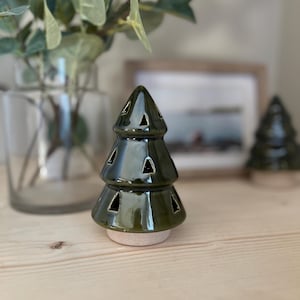 Puede incluir: Un portavelas con forma de árbol de Navidad de cerámica verde oscuro brillante. El árbol tiene cortes triangulares y se apoya sobre una base de color claro. El fondo incluye un jarrón con vegetación y una imagen enmarcada.