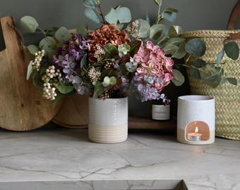 Könnte beinhalten: Blumenarrangement in einer weiß-beigen Vase mit rosa, lila und orangefarbenen Blüten und grünen Blättern. Eine brennende Kerze befindet sich in einem weiß-beigen Keramik-Duftlampen. Ein Holzbrett und ein geflochtener Korb im Hintergrund.