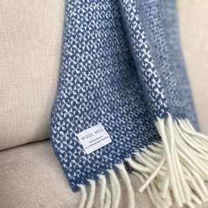 K&ouml;nnte beinhalten: Eine blaue und wei&szlig;e gemusterte Wolldecke mit wei&szlig;en Fransen. Die Decke hat ein Etikett mit der Aufschrift "Wool Hill Made in Britain Cold Wash or Hand Wash/Woolcare"