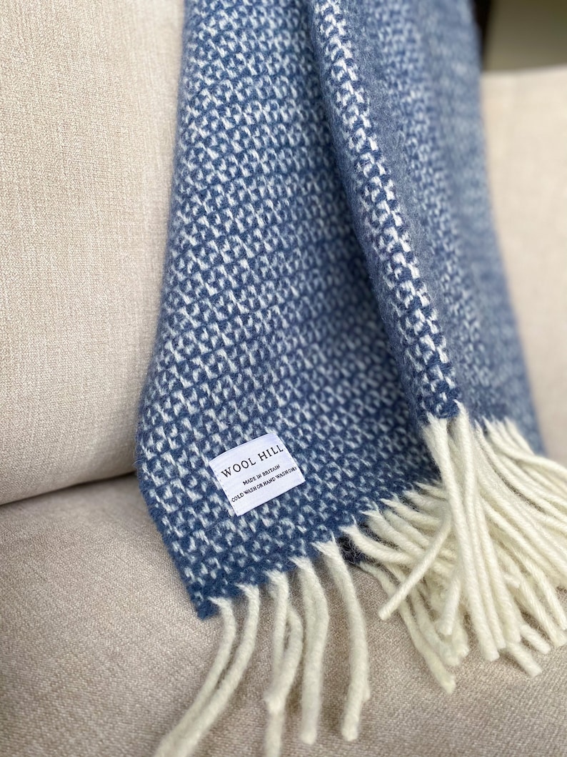 K&ouml;nnte beinhalten: Eine blaue und wei&szlig;e gemusterte Wolldecke mit wei&szlig;en Fransen. Die Decke hat ein Etikett mit der Aufschrift "Wool Hill Made in Britain Cold Wash or Hand Wash/Woolcare"