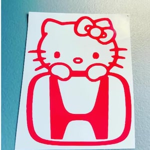 May include: A bright red vinyl decal sticker featuring the iconic Hello Kitty character with a bow, positioned directly above and integrated into the center of a bold red Honda automotive logo emblem on a plain white rectangular backing sheet.