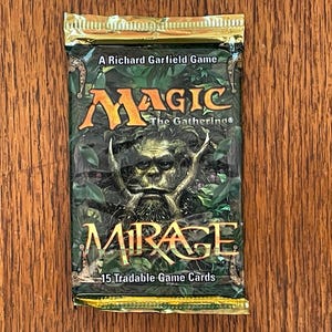 May include: A sealed Magic The Gathering Mirage booster pack featuring a green and brown forest illustration with a central creature face. The text reads A Richard Garfield Game, Magic The Gathering, Mirage, and 15 Tradable Game Cards.