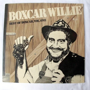 May include: Album cover for "Boxcar Willie: Best of Boxcar, Vol. One." The cover features a black and white illustration of a smiling man in a hat and overalls, holding a bandana. The background is a wooden door with the album title in large, bold letters.