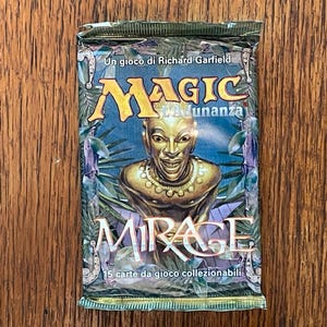 May include: A sealed Magic: The Gathering Mirage booster pack featuring a gold-colored bust illustration on a blue and green background. The packaging displays the text Un gioco di Richard Garfield, Magic L'Adunanza, Mirage, and 15 carte da gioco collezionabili.
