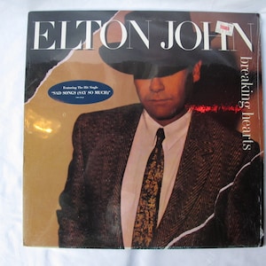 May include: Elton John's "Breaking Hearts" album cover. The image features Elton John in a suit and hat. The album title and artist name are prominently displayed. A sticker is visible in the upper right corner. The album cover also features the text "Sad Songs (Say So Much)".