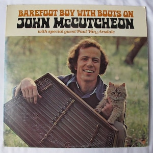 May include: Album cover for "Barefoot Boy With Boots On" by John McCutcheon, featuring a smiling man holding a dulcimer and a cat. The album title is in orange and black text.
