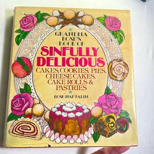 May include: Vintage cookbook titled "Grandma Rose's Book of Sinfully Delicious" featuring illustrations of cakes, cookies, and pastries. The cover is adorned with pink roses, peaches, and baked goods, with text in a decorative font.