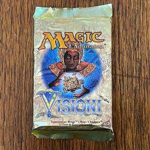 May include: A sealed gold-colored foil booster pack for the Magic: The Gathering card game, specifically the Visioni expansion. The packaging features an illustration of a figure holding a mechanical object, with the text Magic L'Adunanza and Visioni.