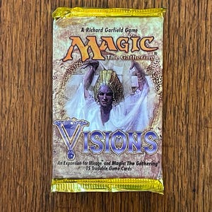 May include: A sealed Magic The Gathering Visions expansion booster pack with gold foil edges. The packaging features a central illustration of a figure in white robes and a gold headdress, with the text A Richard Garfield Game, Magic The Gathering, and Visions.