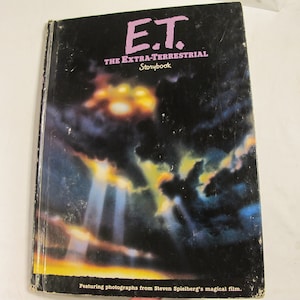 May include: A vintage E.T. The Extra-Terrestrial Storybook. The cover features the film's title in pink and white, set against a dark, space-themed background with a bright light. The book is a collectible item from Steven Spielberg's magical film.