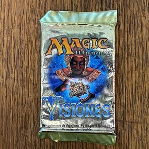 May include: A sealed silver foil booster pack for the Magic: The Gathering card game, specifically the Visions expansion set. The packaging features a central illustration of a figure holding a mechanical object, with the text Magic El Encuentro and Visiones printed in blue and gold lettering.