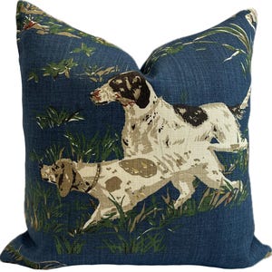 May include: Square cushion with a navy blue background featuring a print of two dogs in a grassy field. The dogs are white with brown and black spots. Green foliage is scattered throughout the design.