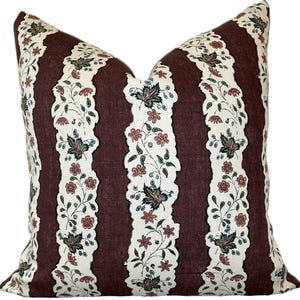 Op de afbeelding: Een decoratief kussen met een streepjespatroon. Afwisselende verticale strepen in donker bordeaux en gebroken wit. De gebroken witte strepen hebben een bloemen- en bladerontwerp in rood, groen en blauw. Het kussen heeft een zachte, getextureerde uitstraling.