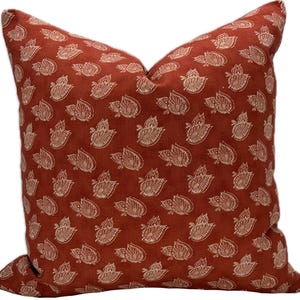 May include: A square decorative pillow with a rust-colored background and a repeating pattern of off-white floral motifs. The pillow is filled and has a soft, inviting appearance, suitable for a living room or bedroom.
