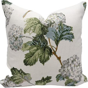 Puede incluir: Un cojín decorativo cuadrado con fondo blanco y estampado floral. El patrón presenta hojas verdes, flores gris-azuladas y tallos marrones. El cojín probablemente está hecho de una tela suave.