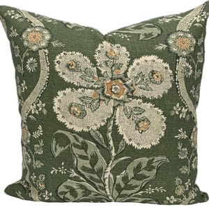 May include: A square throw pillow featuring a dark olive green background with a large, central floral pattern in cream, light green, and muted orange. The design includes intricate stippled textures on the petals and leaves.