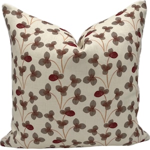 May include: Cream-colored decorative pillow with a repeating pattern of brown and burgundy clover-like shapes. The pillow has a soft, textured appearance, suggesting a linen or similar fabric. The design is symmetrical and botanical in theme.