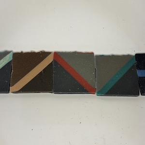 Op de afbeelding: Vijf rechthoekige stukken zeep op een rij. Elk stuk heeft een donkere basiskleur met een contrasterende diagonale streep in lichtblauw, beige, rood, groenblauw, of een horizontale blauwe streep, wat een geometrisch patroon op de set creëert.