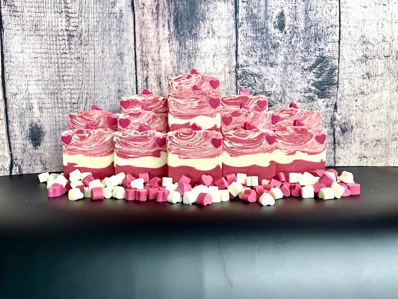 May include: A stack of handmade soap bars with a pink and white swirl design. The bars are decorated with small pink hearts and are surrounded by a scattering of pink and white heart-shaped soap pieces.