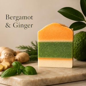 May include: A rectangular bar of soap featuring horizontal layers of orange, green, and cream colors. The text Bergamot & Ginger is printed in dark green on the left side. The soap is positioned near fresh ginger root and a green citrus fruit.