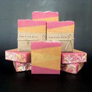 Può includere: Barrette di sapone fatte a mano nei toni del rosa e dell'arancione, impilate e disposte. Le barrette sono a strati con un motivo marmorizzato sul fondo. Alcune barrette sono avvolte con spago e un'etichetta con la scritta "Zion Creek Farm" e "Desert Flower".
