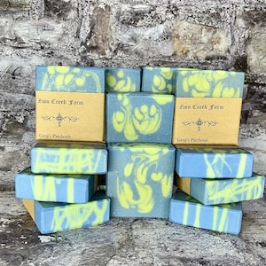 Op de afbeelding: Een stapel handgemaakte zeepjes met een blauw en groen swirl-patroon. De zeepjes zijn gelabeld met "Zion Creek Farm" en "Greg's Patchouli".