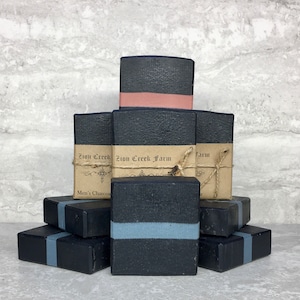 May include: A stack of black charcoal soap bars with a light blue stripe. The top bar has a brown label that reads "Zion Creek Farm".