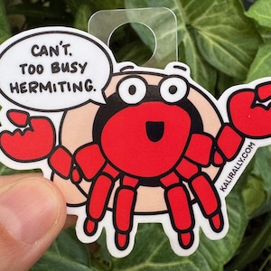 May include: A red cartoon hermit crab with a speech bubble that says "Can't. Too busy hermitiing." The crab is holding a shell and has a happy expression on its face. The sticker is on a green leafy background.